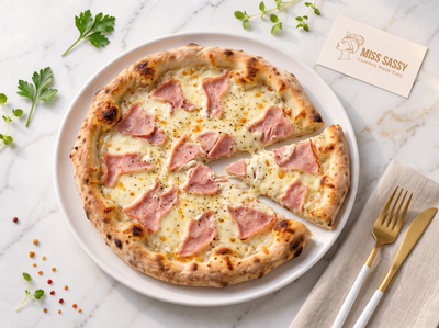 A freshly baked pizza with ham and melted cheese on a white plate, styled on a marble surface with parsley, peppercorns, a gold fork, a knife, and a card reading Miss Sassy.