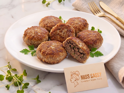Six browned meatballs on a white plate, with one cut open to show the textured interior. The plate is set on a marble surface with a beige napkin, gold-toned fork and knife, and a small card reading MISS SASSY Comfort Made Easy.