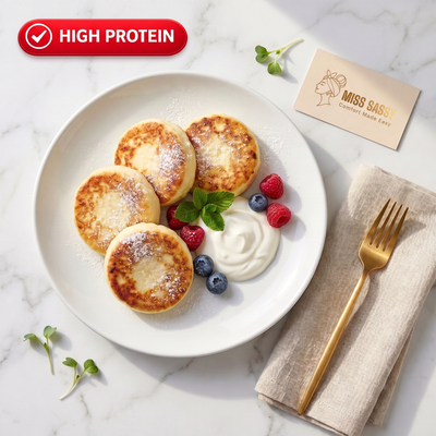 Four golden-brown pancakes on a white plate with a dollop of whipped cream, blueberries, raspberries, and mint, styled on a white marble surface with a gold fork, a beige napkin, and a Miss Sassy card. A red badge at the top left reads 'HIGH PROTEIN'.